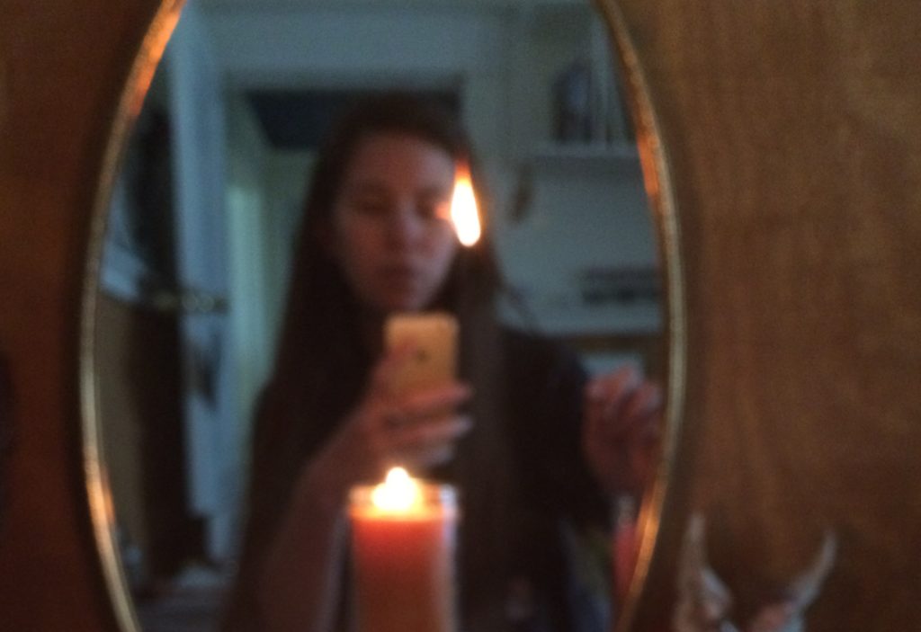 a blurry photo of Paige reflected in an oval mirror. She is taking a selfie in the mirror and holding a lit stick with a large flame at the end. A large lit candle stands before her. The light from the flames glints off the gold frame of the mirror.